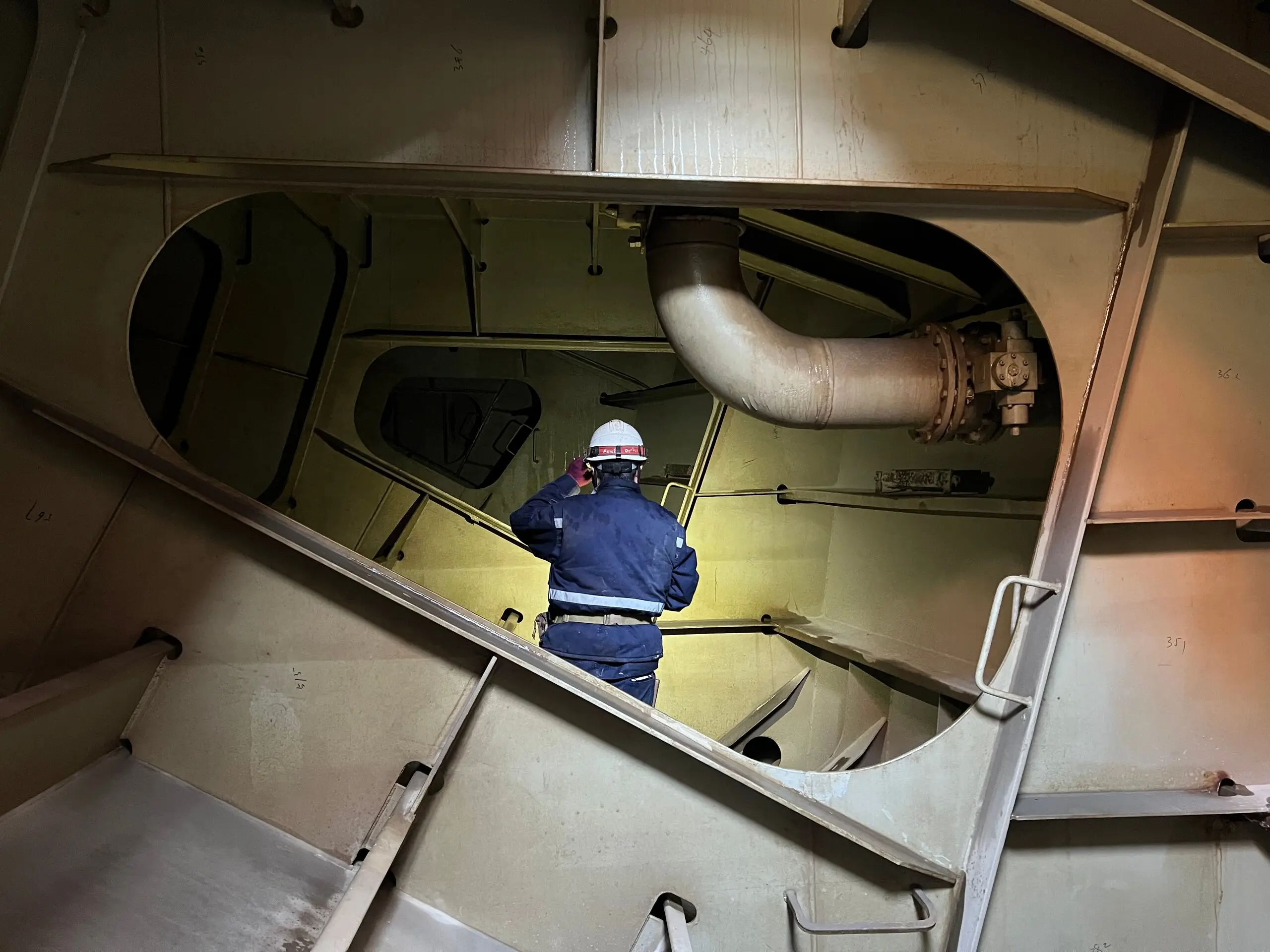 Ship tank interior seen through an oval manhole, a crew member in overalls and a white helmet standing inside.